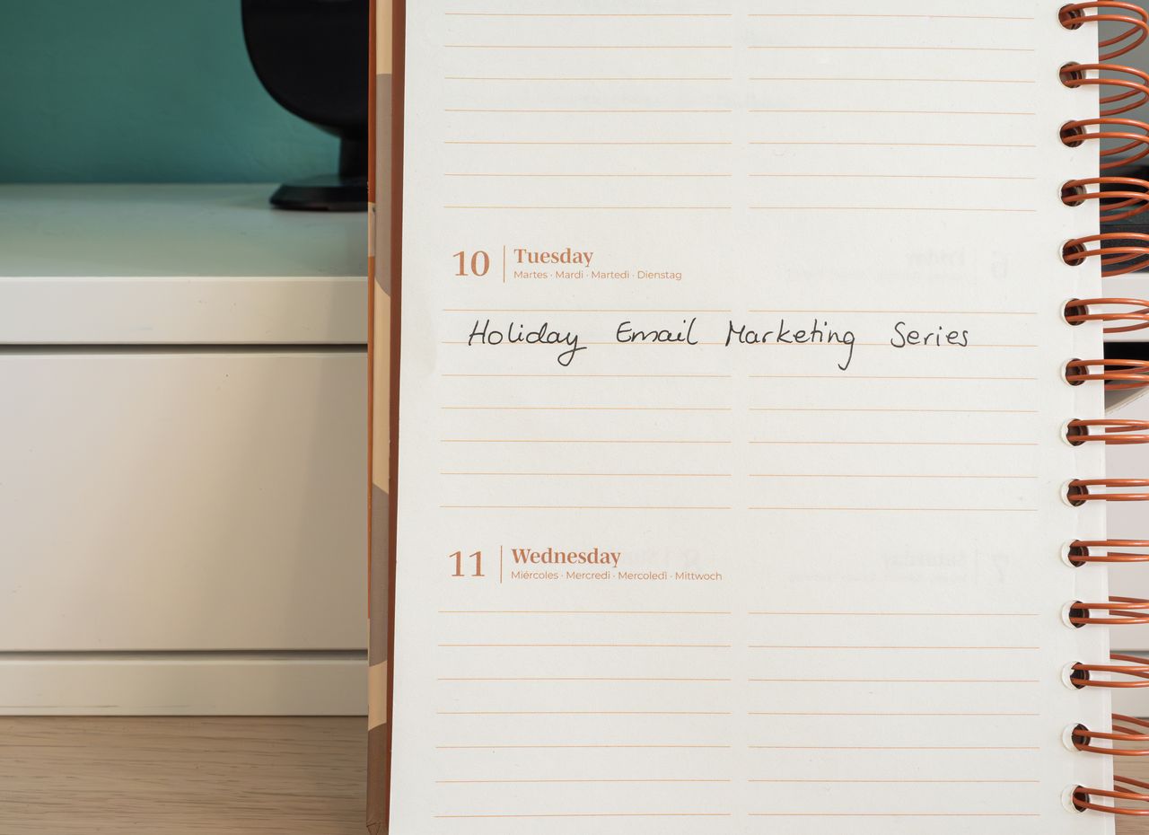 A planner lies open on a desk beside a handwritten note for a Holiday Email Marketing Series.